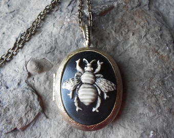 May include: Oval-shaped pendant necklace with a black background and a cream-colored bee design. The pendant is framed in a gold-toned metal with an ornate border and hangs from a matching chain. The necklace is a vintage-inspired accessory.