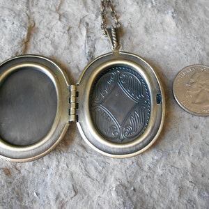 Pode incluir: Um colar com um medalh&atilde;o oval, com um design decorativo na parte de tr&aacute;s. O medalh&atilde;o est&aacute; aberto, revelando um interior em branco. O medalh&atilde;o &eacute; aproximadamente do tamanho de uma moeda de 25 centavos.