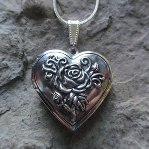 May include: A silver heart-shaped locket pendant with a rose design. The pendant hangs from a silver chain. The locket is detailed with an embossed rose and leaf pattern. The background is a textured gray stone.