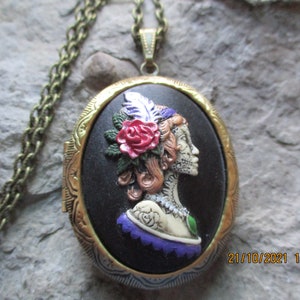May include: A gold locket necklace with a black oval cameo featuring a sugar skull woman with a red rose and feather in her hair. The cameo is surrounded by a gold filigree design.