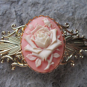 May include: A gold-tone hair clip with a cameo featuring a white rose on a pink background.