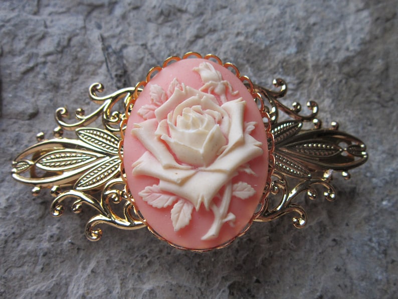 May include: A gold-tone hair clip with a cameo featuring a white rose on a pink background.