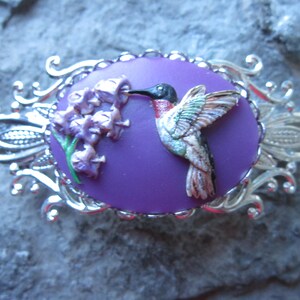 May include: A silver-toned brooch with a purple cameo featuring a hummingbird in flight, drinking nectar from a cluster of purple flowers.