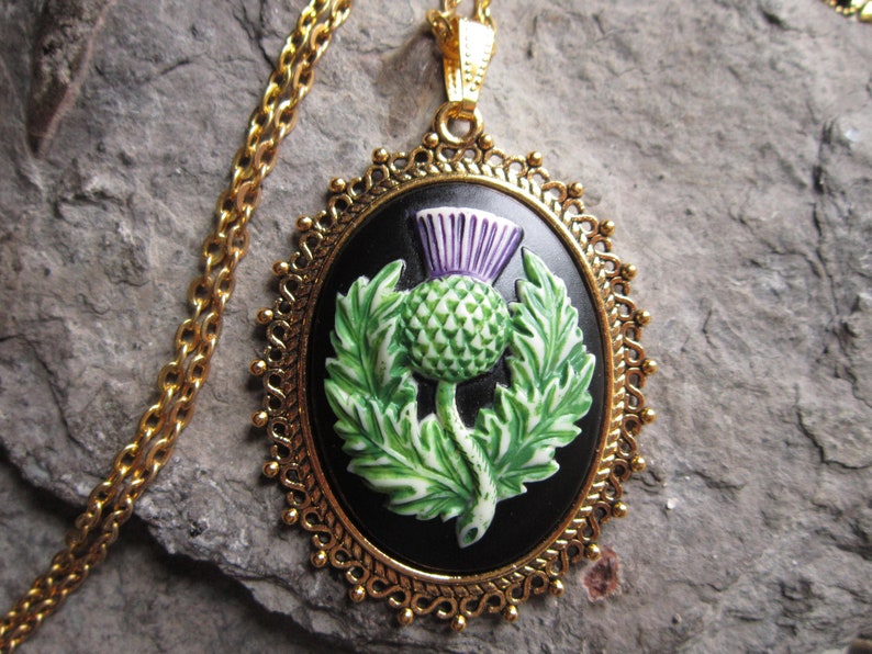 May include: Gold-toned necklace with an oval pendant featuring a black background and a detailed thistle design in green, white, and purple. The pendant is framed by a decorative gold border.