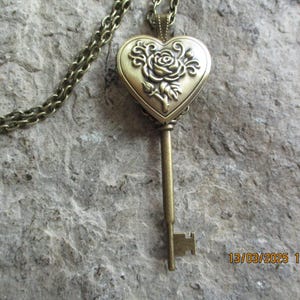 May include: A vintage-style necklace featuring a bronze-colored key pendant. The pendant has a heart-shaped locket with a rose design. The key is suspended from a matching chain, creating a romantic and antique aesthetic.