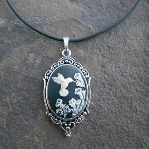 May include: A black cameo pendant necklace with a white hummingbird and flowers design. The pendant is set in a silver-toned ornate frame and hangs from a black cord.