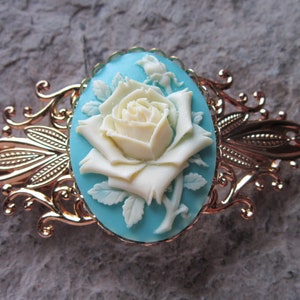 May include: A gold-tone hair clip with a decorative design features a white cameo of a rose on a turquoise background.