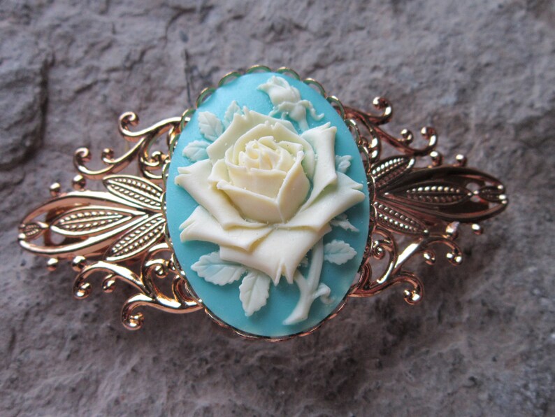 May include: A gold-tone hair clip with a decorative design features a white cameo of a rose on a turquoise background.
