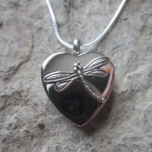 May include: Silver heart-shaped locket necklace with a dragonfly design. The locket is open and the inside is visible.