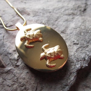 May include: A gold oval pendant necklace with two detailed turtle charms. The pendant is on a gold chain.