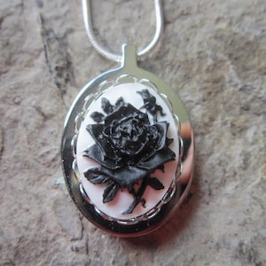 Può includere: Una collana con un ciondolo d'argento con un cammeo nero che presenta una singola rosa nera su uno sfondo bianco.