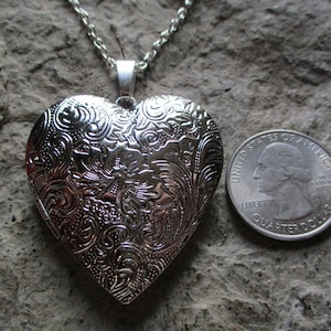 May include: A silver heart-shaped locket necklace with intricate floral and scrollwork detailing. The locket is open and has a chain. A U.S. quarter is next to the locket for size comparison. The locket is on a textured, gray surface.