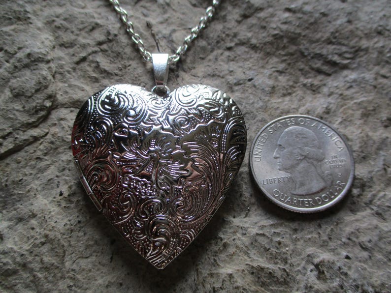 May include: A silver heart-shaped locket necklace with intricate floral and scrollwork detailing. The locket is open and has a chain. A U.S. quarter is next to the locket for size comparison. The locket is on a textured, gray surface.