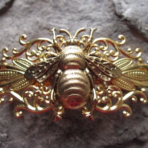 May include: A gold-toned bee brooch with intricate filigree details. The bee is centred on a decorative, ornate frame.