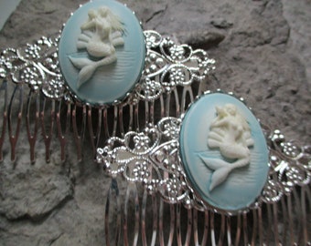 May include: Two silver hair combs with blue and white cameo mermaid designs. The mermaids are in a reclining pose with their tails flowing behind them.