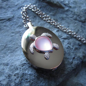 May include: A silver chain necklace with a silver oval locket featuring a pink sea turtle charm.