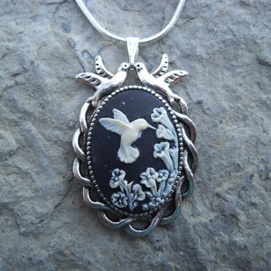 May include: Silver-toned pendant necklace featuring an oval cameo with a white hummingbird and flowers on a black background. The pendant is framed with a silver rope-like border and topped with two silver bird figures.