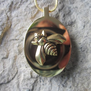 Puede incluir: Un collar con un colgante de abeja de oro. La abeja está en relieve y tiene un cuerpo texturizado. El colgante es de forma ovalada y cuelga de una cadena de oro.