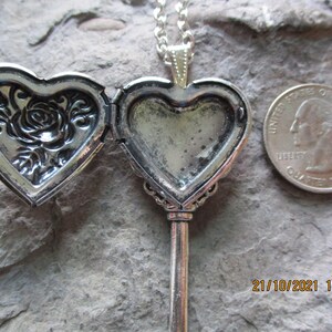 May include: Silver-toned heart-shaped locket necklace with a key design. The locket opens to reveal space for a photo. One side features a rose design. A U.S. quarter is next to the locket for size comparison.