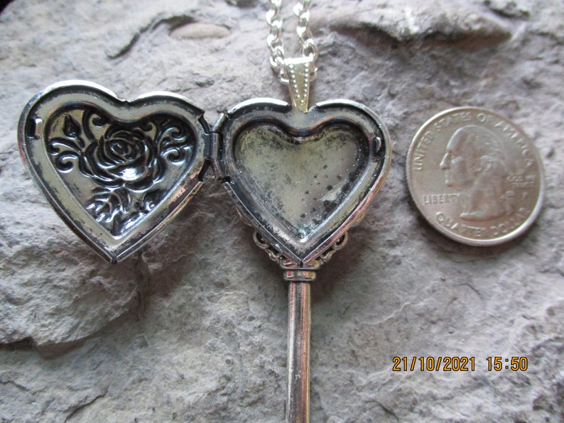 May include: Silver-toned heart-shaped locket necklace with a key design. The locket opens to reveal space for a photo. One side features a rose design. A U.S. quarter is next to the locket for size comparison.