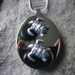 May include: A silver pendant necklace featuring two detailed turtle silhouettes. The pendant is oval shaped and has a shiny, reflective surface.