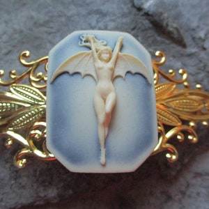 Puede incluir: Una pinza para el pelo de metal dorado con un camafeo azul y blanco que representa a una mujer desnuda con alas de murciélago.