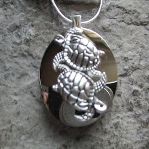 May include: A silver pendant necklace featuring two detailed turtle charms. The turtles are intertwined and have a textured shell design. The pendant is attached to a silver chain.