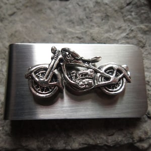 Puede incluir: Un clip de dinero de metal plateado con un diseño detallado de motocicleta. La motocicleta es de estilo crucero clásico, con ruedas, manillar y componentes del motor visibles. El clip tiene un acabado de metal cepillado.