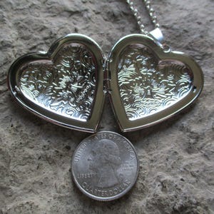 May include: Open silver heart-shaped locket with ornate floral detailing. A U.S. quarter coin is placed below the locket for size comparison. The locket is on a textured, gray surface. The locket is attached to a silver chain.