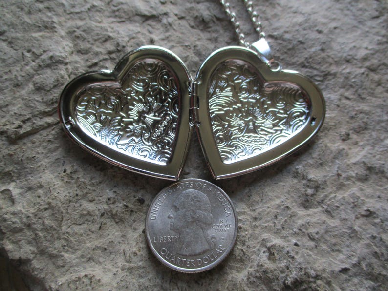 May include: Open silver heart-shaped locket with ornate floral detailing. A U.S. quarter coin is placed below the locket for size comparison. The locket is on a textured, gray surface. The locket is attached to a silver chain.