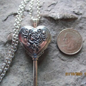 May include: Silver-toned heart-shaped locket pendant necklace with a rose design. The pendant is key-shaped and has a chain. A U.S. quarter is next to the pendant for size comparison.