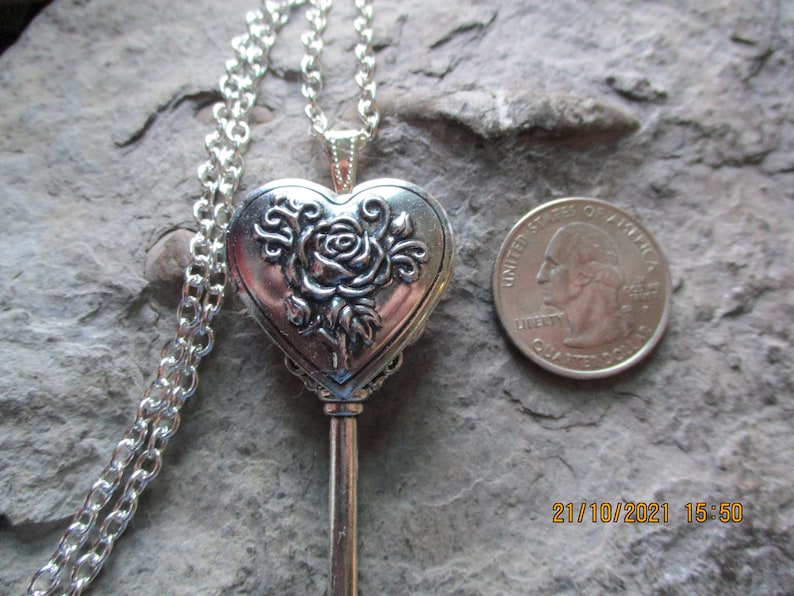 May include: Silver-toned heart-shaped locket pendant necklace with a rose design. The pendant is key-shaped and has a chain. A U.S. quarter is next to the pendant for size comparison.
