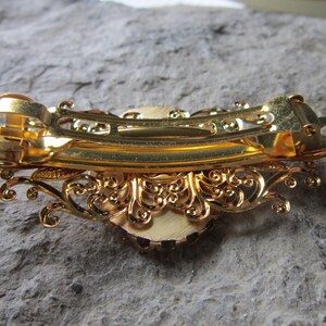 May include: Gold hair clip with a decorative design and a large, round, faux gem in the center.