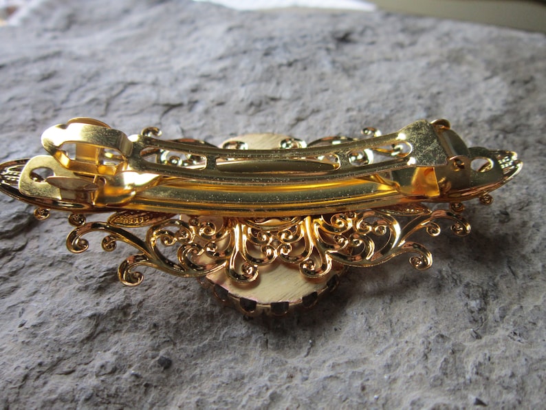 May include: Gold hair clip with a decorative design and a large, round, faux gem in the center.