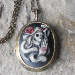 May include: A silver chain necklace with a bronze oval locket featuring a black background with a white skull and a snake, a red rose, and a red heart.