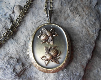 May include: A gold-toned oval locket necklace with a chain. The locket features two detailed bee designs.