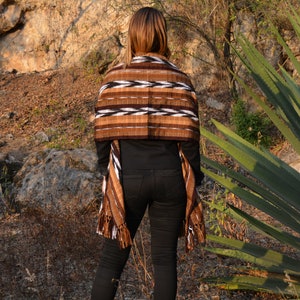 May include: A woman wearing a brown, white and black woven shawl with fringe. The shawl has a geometric pattern. She is standing in front of a large green plant with long, sharp leaves.