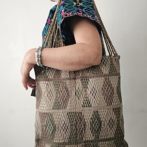 Puede incluir: Un bolso tote tejido con un patrón geométrico en tonos marrones y verdes. El bolso tiene asas trenzadas y lo lleva una persona que viste una blusa bordada colorida. El diseño del bolso presenta formas de diamante y rectangulares.