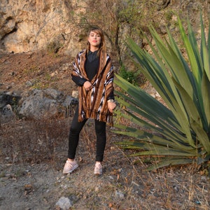 May include: A woman wearing a brown and white patterned shawl with black trousers and pink trainers. She is standing in front of a large green plant and a rocky background.