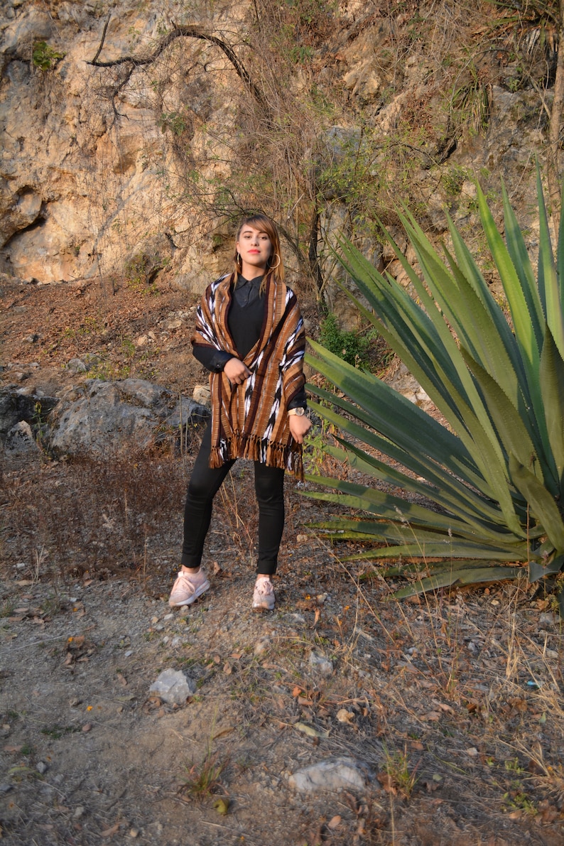 May include: A woman wearing a brown and white patterned shawl with black trousers and pink trainers. She is standing in front of a large green plant and a rocky background.