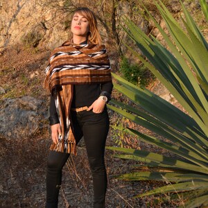 May include: A woman wearing a brown and white patterned shawl with black trousers and pink trainers. She is standing in a field with a large green plant behind her.