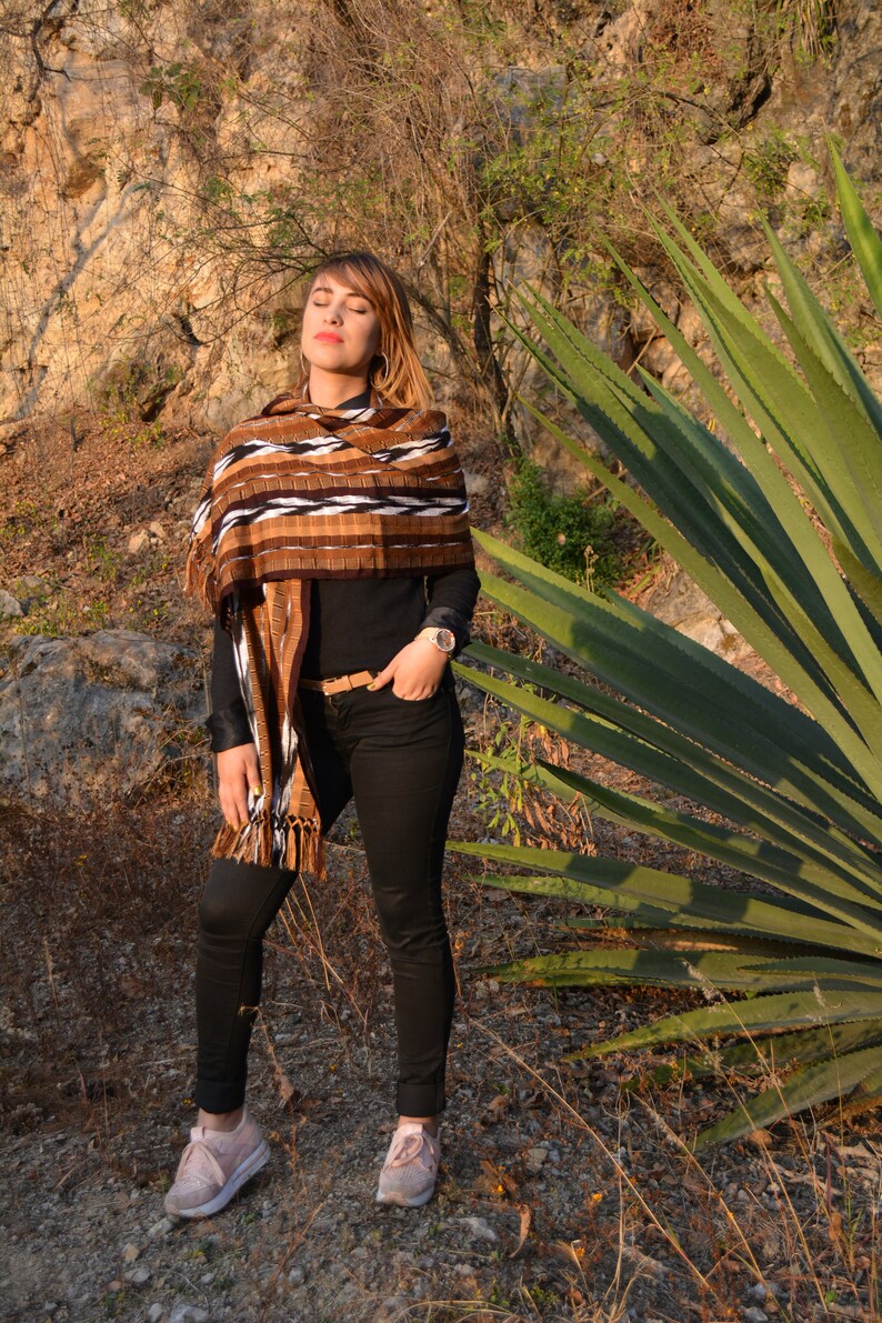 May include: A woman wearing a brown and white patterned shawl with black trousers and pink trainers. She is standing in a field with a large green plant behind her.
