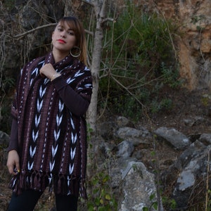 May include: A woman wearing a brown and white patterned shawl with fringe. The shawl is woven with a geometric design. She is standing in front of a rocky background.