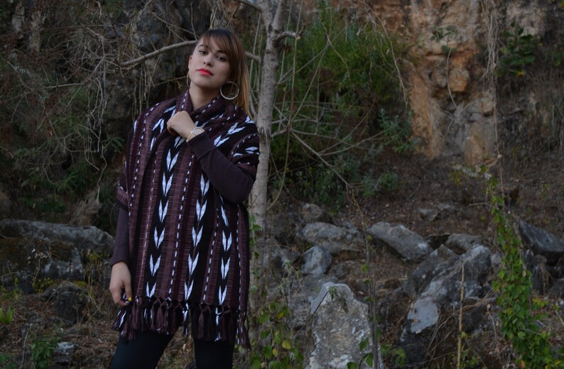 May include: A woman wearing a brown and white patterned shawl with fringe. The shawl is woven with a geometric design. She is standing in front of a rocky background.