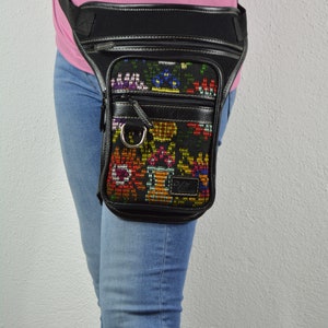 May include: A black leather fanny pack with a colorful embroidered design. The fanny pack is worn on the hip of a person wearing blue jeans.