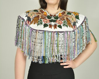 May include: A white embroidered top with a colorful bird and floral design. The top features a fringed hem with strands of various colors. The model is wearing black leggings and a patterned scrunchie.