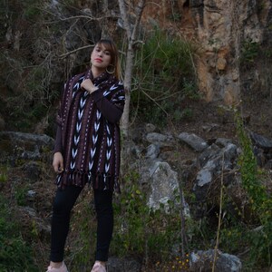May include: A woman wearing a brown and white patterned poncho with black trousers and pink trainers. She is standing on a large rock in a rocky, outdoor setting.