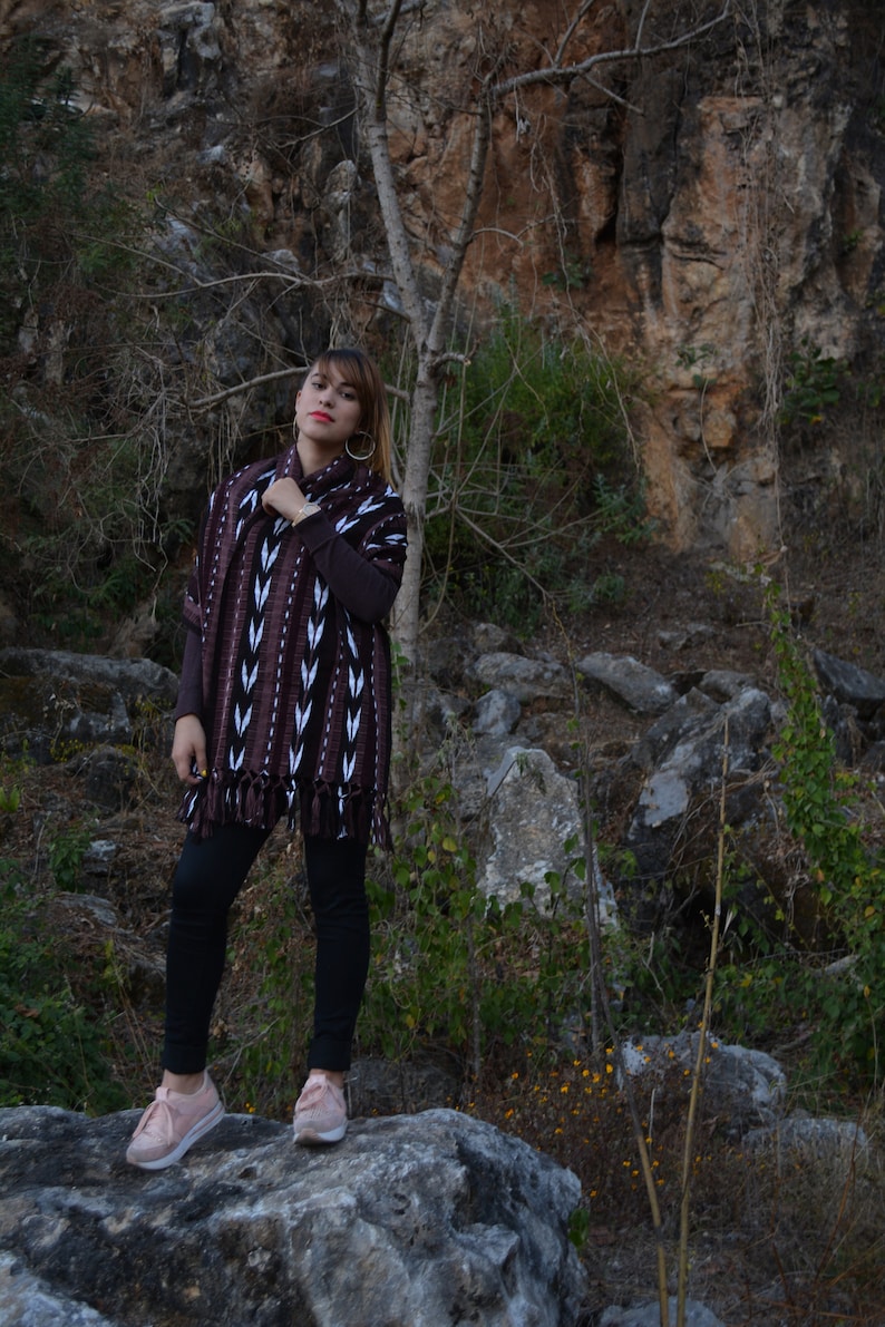 May include: A woman wearing a brown and white patterned poncho with black trousers and pink trainers. She is standing on a large rock in a rocky, outdoor setting.