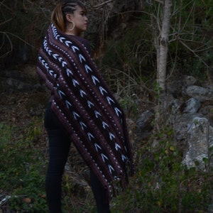 May include: A woman wearing a brown and white patterned shawl with fringe. She is standing on a rocky surface with green foliage in the background.