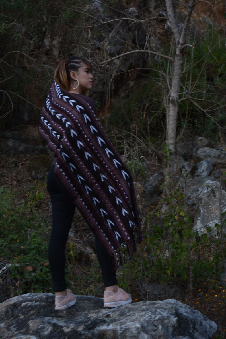 May include: A woman wearing a brown and white patterned shawl with fringe. She is standing on a rocky surface with green foliage in the background.